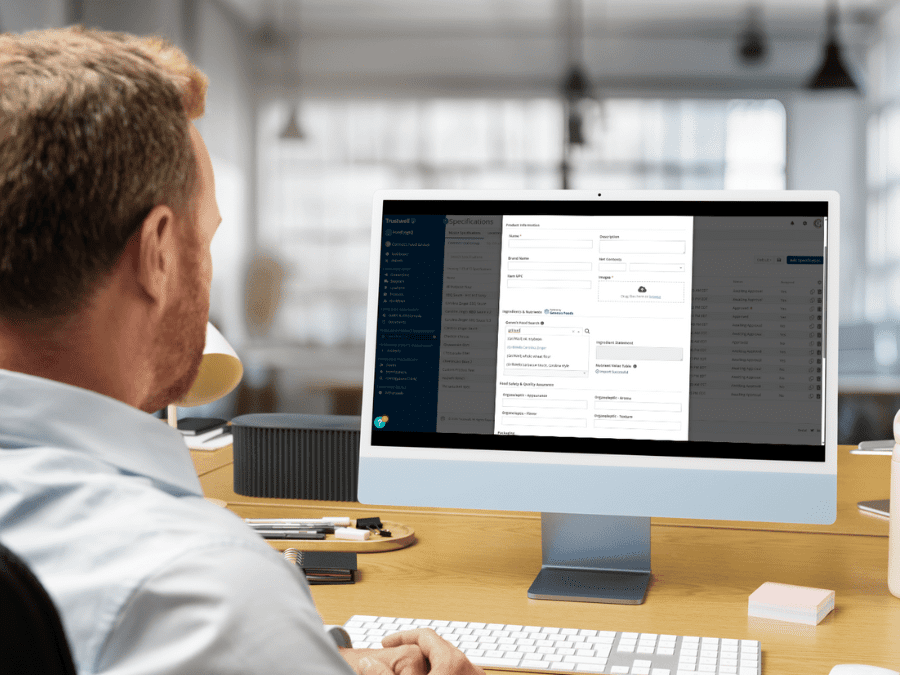 A blond, bearded man sitting at an office desk with a computer screen showing the Add New Spec screen of FoodLogiQ Product Management